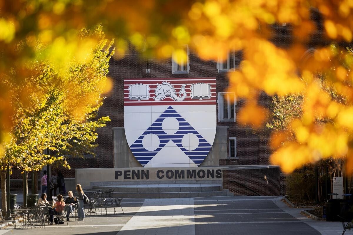 Students beneath the large sculpture of the Penn shield at Penn Commons, framed by brilliant fall foliage.